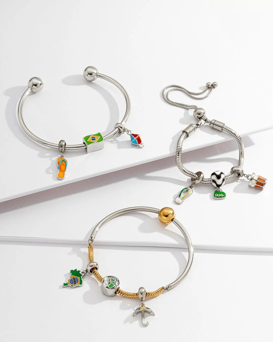 Três pulseiras de berloques prateadas e douradas com pingentes temáticos sobre fundo branco.