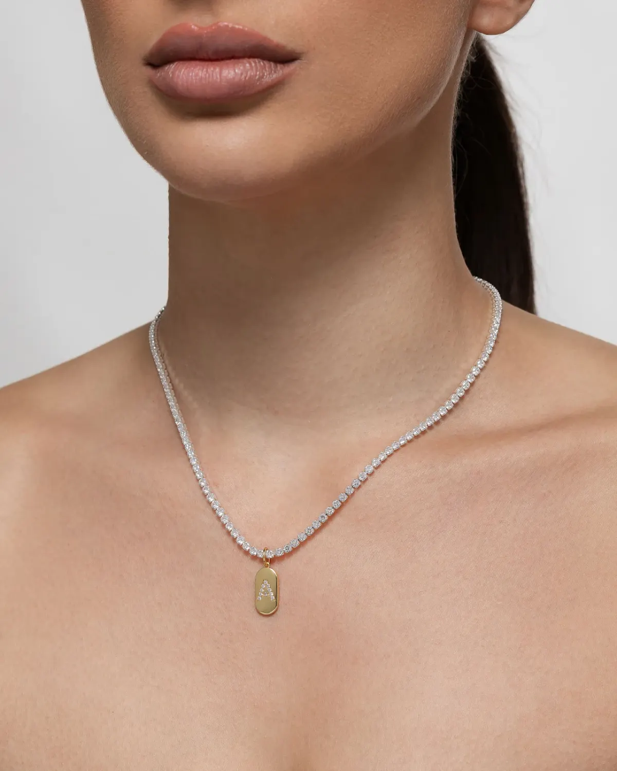 Modelo feminina usando corrente estilo riviera prateada com pingente dourado oval da letra A cravejada.