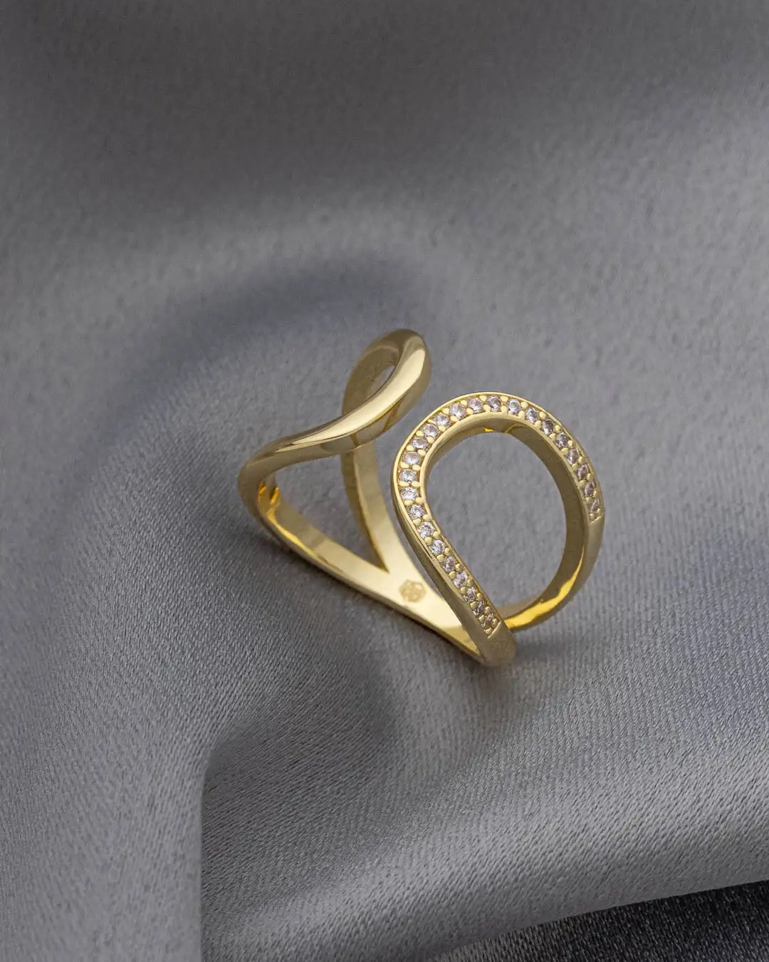 Anel aberto folheado a ouro com design sinuoso e detalhe cravejado de zircônias sobre tecido cinza.